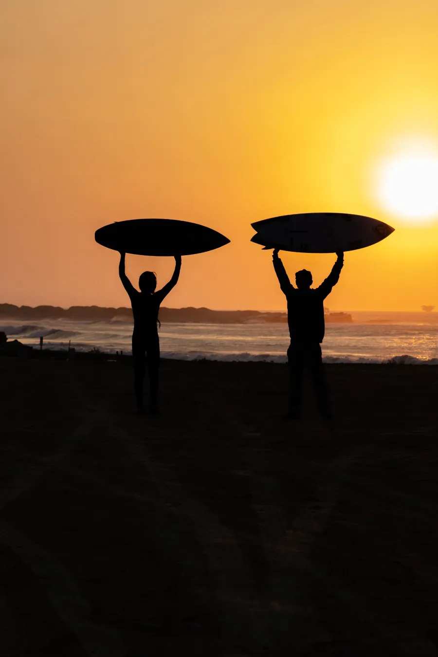 Dois surfistas em silhueta segurando as pranchas acima da cabeça, contra o céu laranja do pôr do sol.